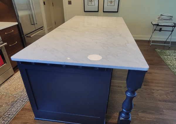 Blue kitchen island with a white marble top and a turned leg, in a home kitchen with stainless appliances nearby.